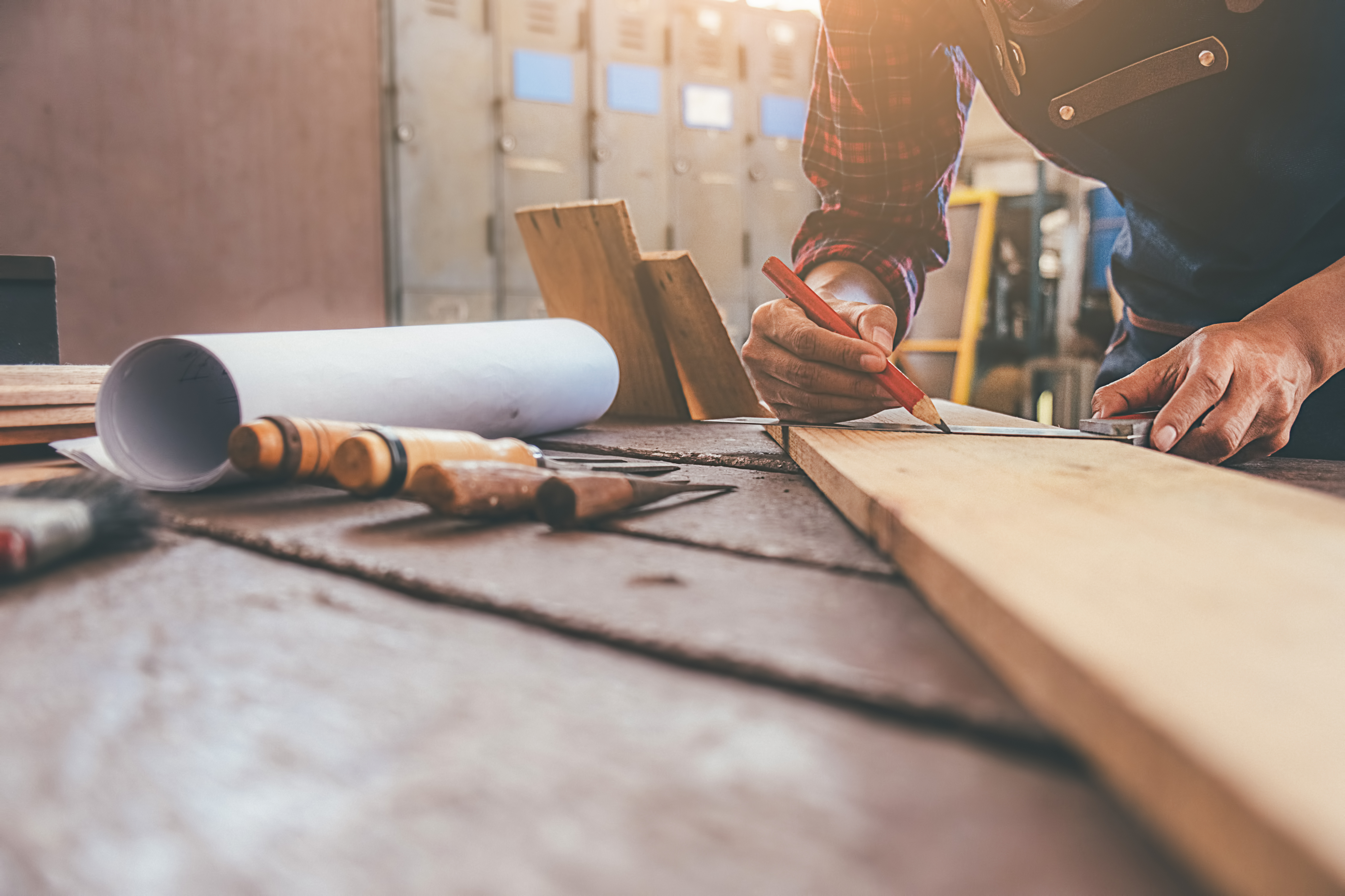Handwerker markiert Messpunkte auf einem Holzbrett auf einer Werkbank mit Werkzeugen und Bauplänen im Vordergrund.