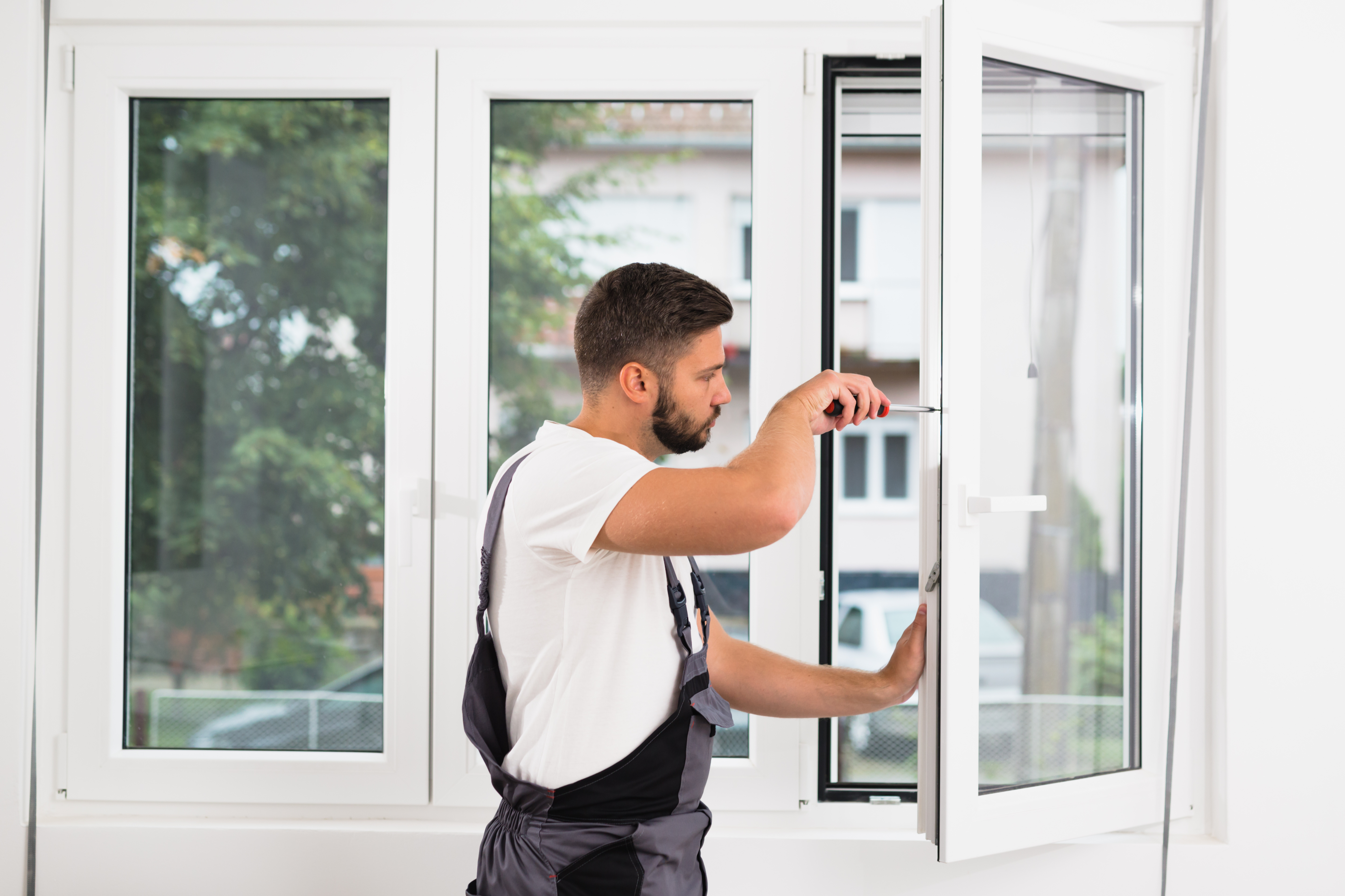 Handwerker im weißen Hemd und mit Hosenträgern justiert ein Fenster in einem hellen Raum. Er konzentriert sich auf die Feineinstellung des Fenstergriffs.