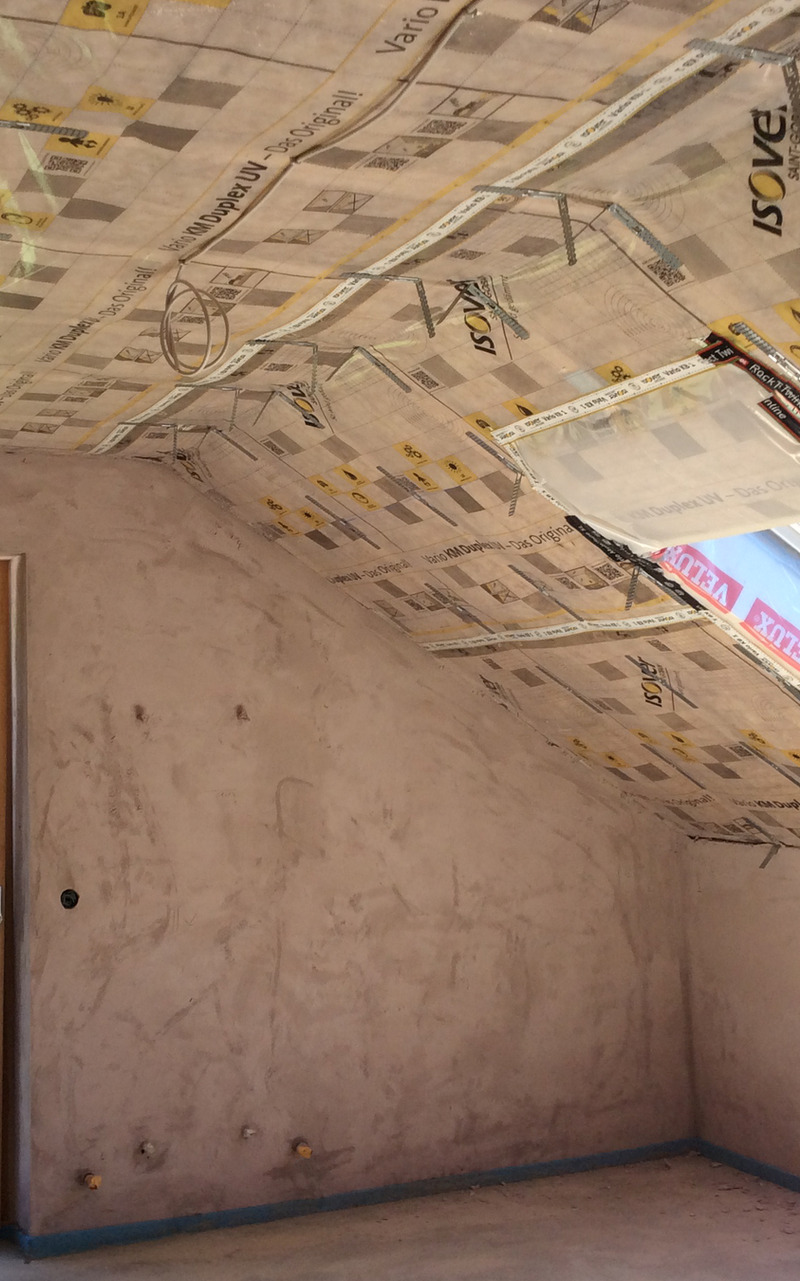 Ecke eines Raumes im Bau mit Dampfsperrfolie an der Dachschräge und sichtbaren Gipskartonplatten an den Wänden. Die Folie trägt Aufdrucke und Markierungen, die auf die Isolierung hinweisen.